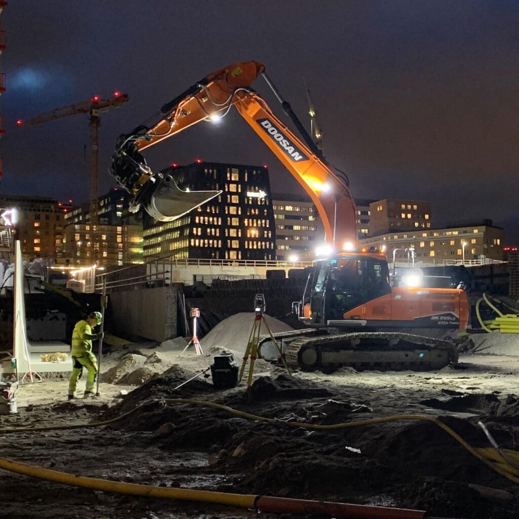 En Doosan grävmaskin från Vabeko arbetar på en upplyst byggarbetsplats i stadsmiljö under natten.